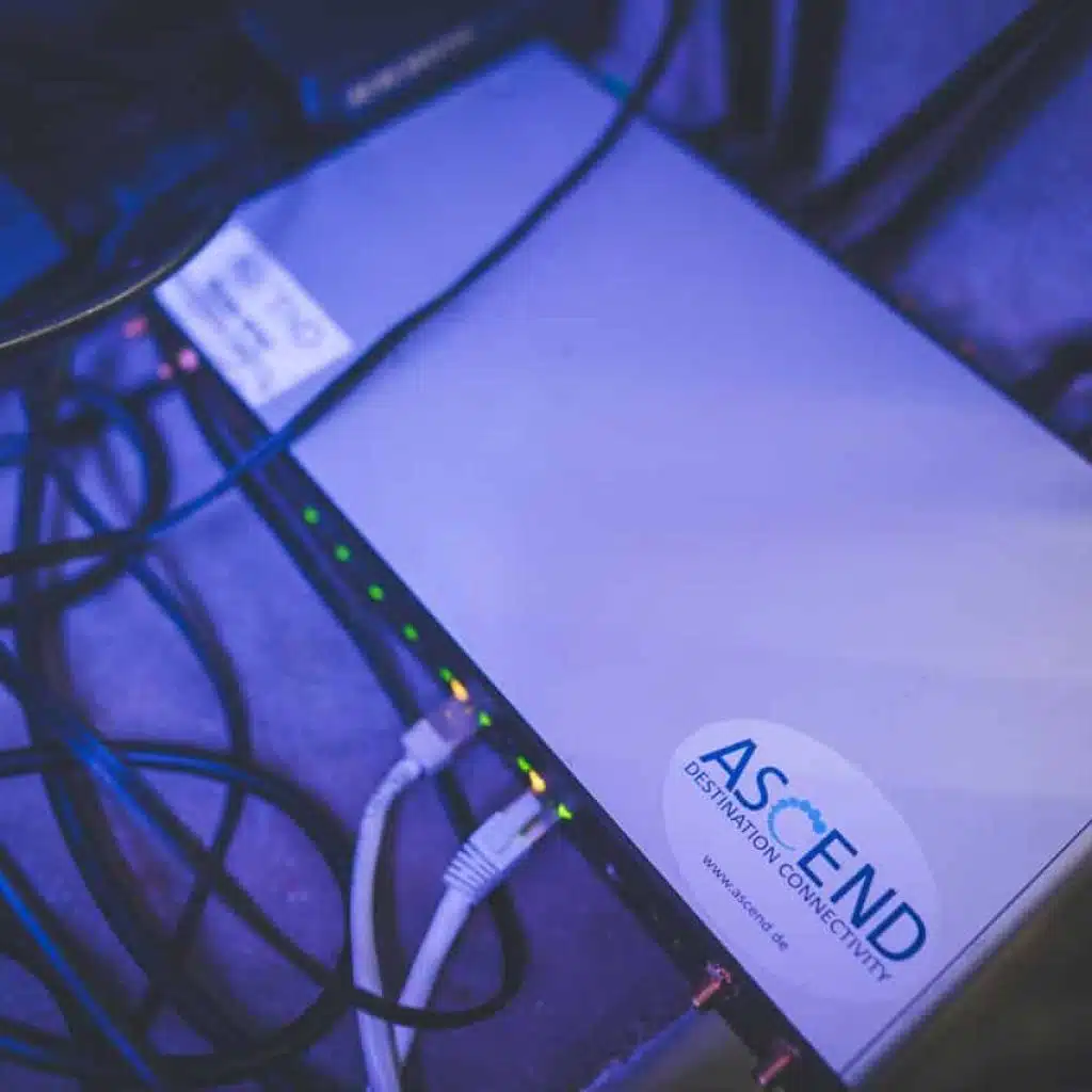 Mobile router from Peplink, model HD4, with three SIM cards inserted and connected. The device has mobile phone antennas and WiFi antennas, both of which are connected. A sticker with the ASCEND logo is clearly visible on the device.